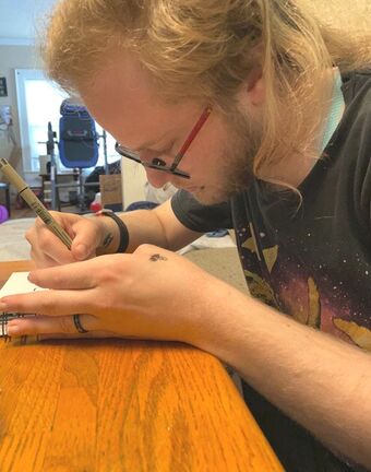 Picture of a long haired blond person with a beard and glasses leaned over a sketchbook and drawing with a pen.